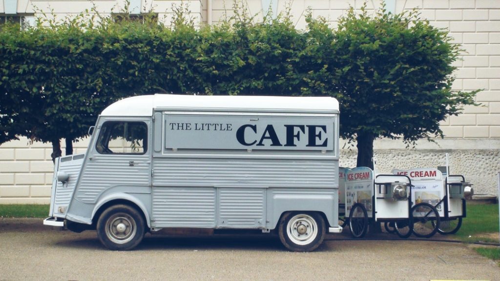 small gray vintage food truck named "the little cafe" parked in front of a row and trees and some ice cream carts