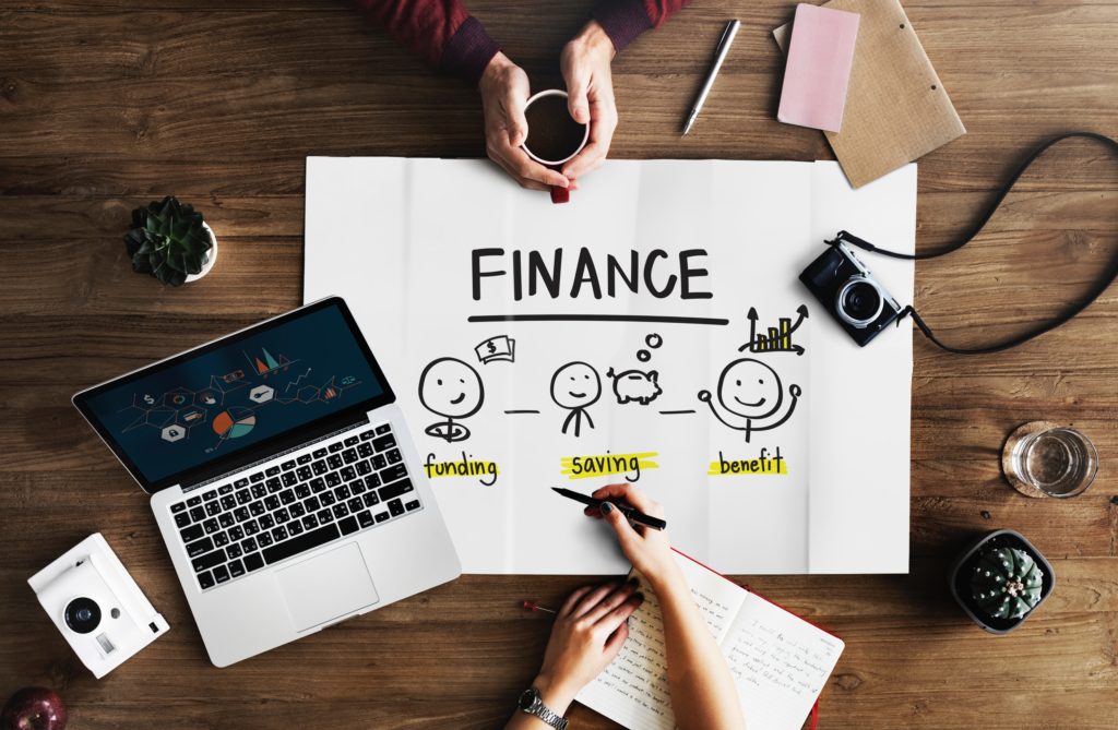 top down view of a wooden desk with one person drawing on a sign that says "finance, funding, saving, benefit" and the other person holding a cup of coffee. Sign is surrounded by a vintage camera, an apple laptop, some notebooks and some succulents.