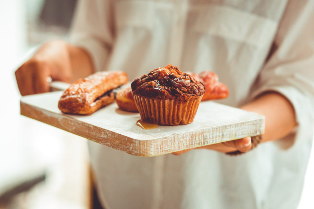 baker holding a wooden serving board with muffin and other pastries on top