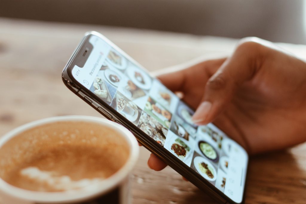 close up photo of a smart phone with the user scrolling through food marketing photos on social media with a cup of coffee in the foreground and wood table top in the background