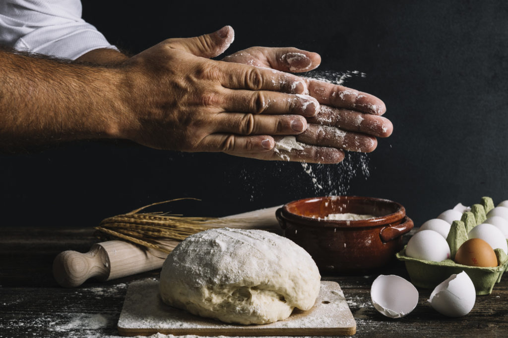 close up of chef hands rubbing flour between them above a ball of dough with various ingredients in the background