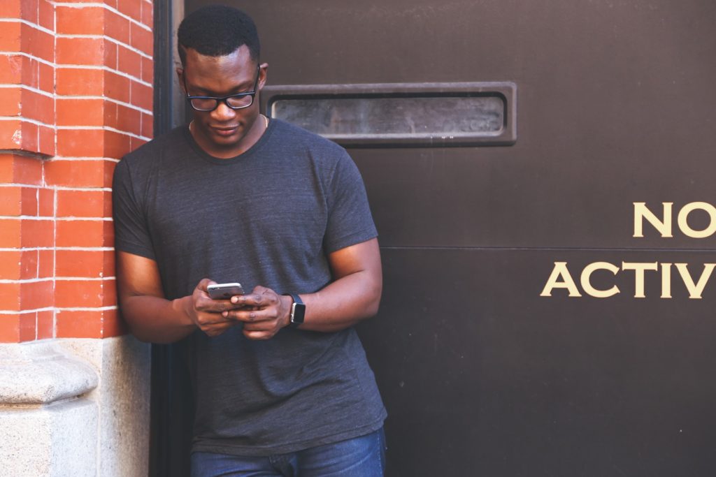 Young black man dressed in clean cut t-shirt and jeans leans against a brick wall looking down at his smartphone