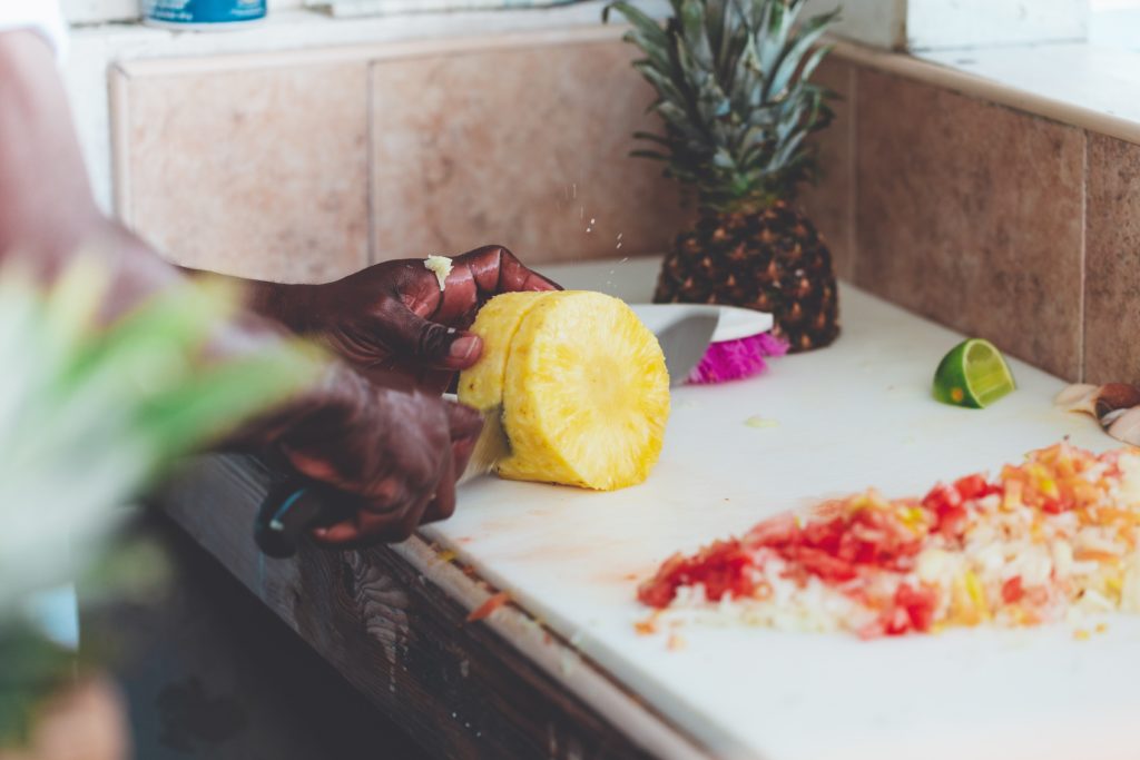 Photograph of black chef's hands cutting up a pineapple with a large knife on a cutting board counter