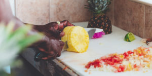 Photo of black chef's hands cutting up a pineapple with large knife on a cutting board counter