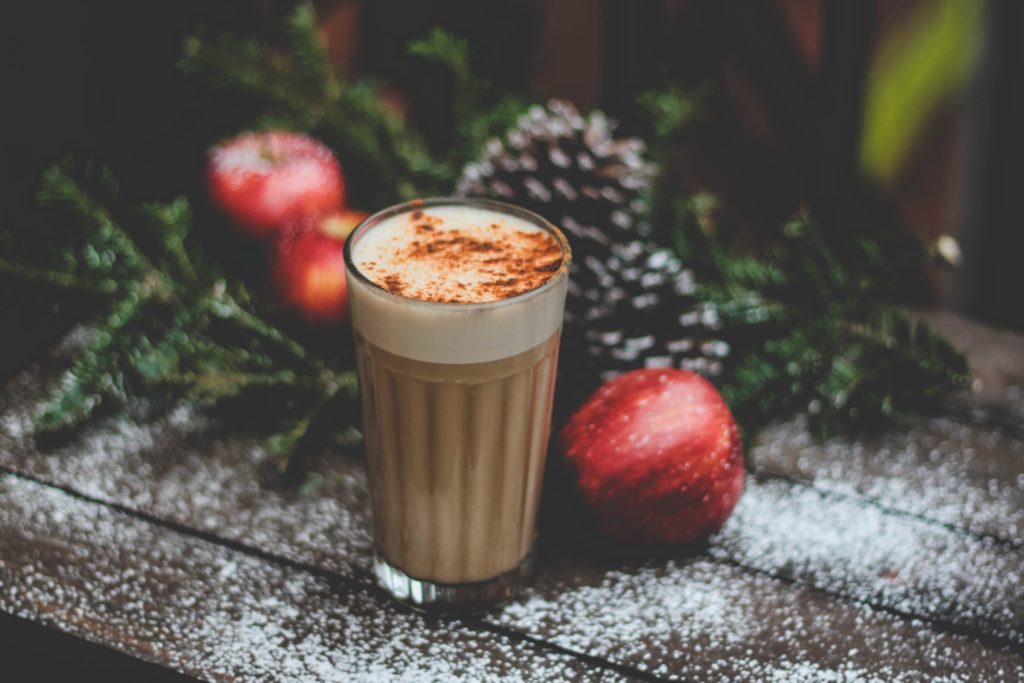 shallow focus photo of a holiday beverage sitting on a snowy bench surrounded by red apples, pine cones and evergreen branches