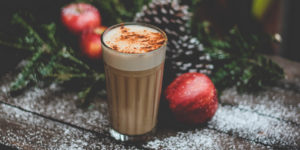 shallow focus photo of a holiday beverage sitting on a snowy bench surrounded by red apples, pine cones and evergreen branches