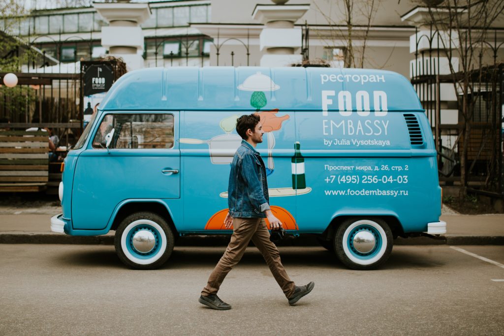 photo of a dark-haired man walking in front of a blue van food truck on a city street