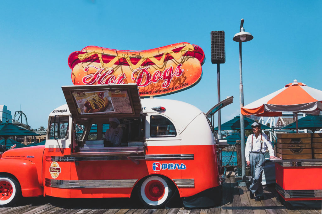 photo of an old fashioned car with a giant hot dog sign on top parked on a pier with an elderly Asian man standing next to it