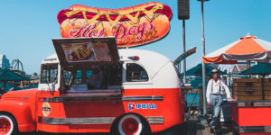 photo of an old fashioned car with a giant hot dog sign on top parked on a pier with an elderly Asian man standing next to it