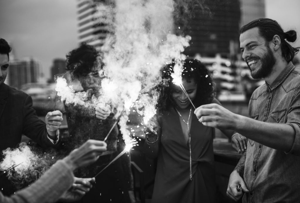 black and photo photo of a diverse group of smiling friends lighting sparklers on a city rooftop with buildings in the background