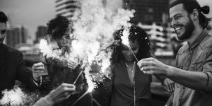 black and photo photo of a diverse group of smiling friends lighting sparklers on a city rooftop with buildings in the background