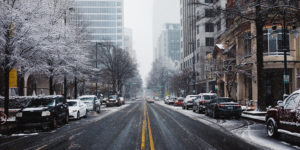 photo of a snowy urban street with trees, parked cars, and buildings