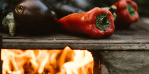 close up photo of red peppers and egg plants being roasted on a grill fire