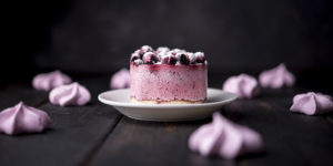 photo of mini pink cheesecake with berries on top on a white plate surrounded by pink frosting puffs sitting on a dark wood table with dark background