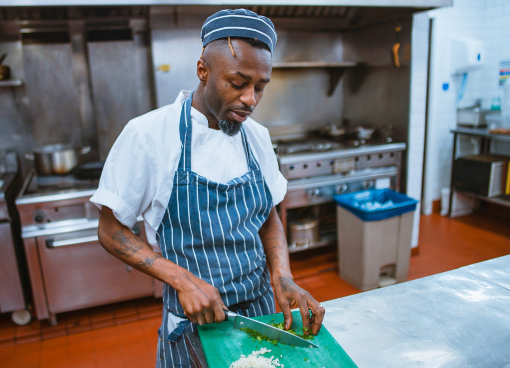 photo of a black man with tattoos on his arms wearing a matching blue-striped apron and chef hat cutting onions in an industrial kitchen setting