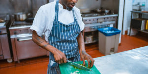 photo of a black man with tattoos on his arms wearing a matching blue-striped apron and chef hat cutting onions in an industrial kitchen setting