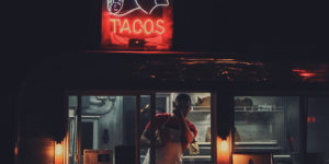 photo at nighttime of a man looking out the window of a food truck with a neon sign on top that says "tacos" with hands holding a taco