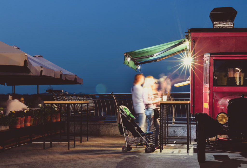 photo of motion-blurred man and woman with a stroller waiting in line under an awning at a red food truck