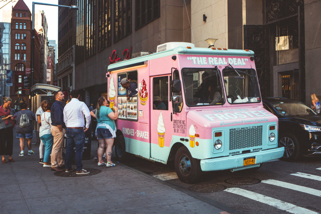 photo of a small pink food truck serving ice cream parked on a city street with a line of customers waiting