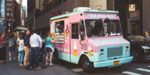 photo of a small pink food truck serving ice cream parked on a city street with a line of customers waiting