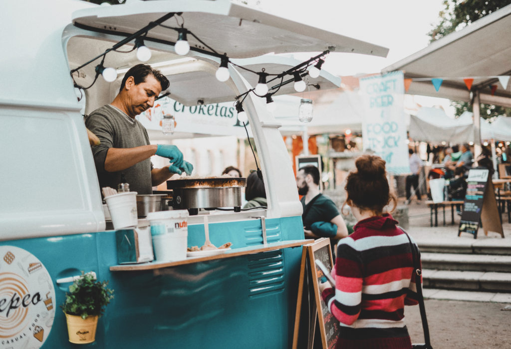 Photo of a woman waiting in front of a vintage blue and white food truck where a man is cooking crepes