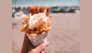 close up of a white person's hand holding a cone of fish and chips with a blurry beach in the background