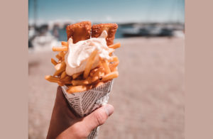 close up of a white person's hand holding a cone of fish and chips with a blurry beach in the background