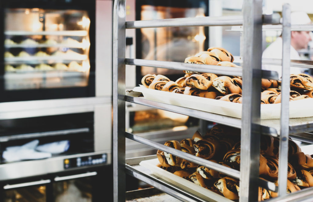 photo of a bakery interior with cinnamon rolls cooling on a baking rack in the foreground and ovens filled with baking goods in the background