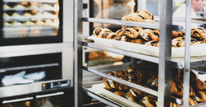 photo of a bakery interior with cinnamon rolls cooling on a baking rack in the foreground and ovens filled with baking goods in the background