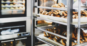 photo of a bakery interior with cinnamon rolls cooling on a baking rack in the foreground and ovens filled with baking goods in the background