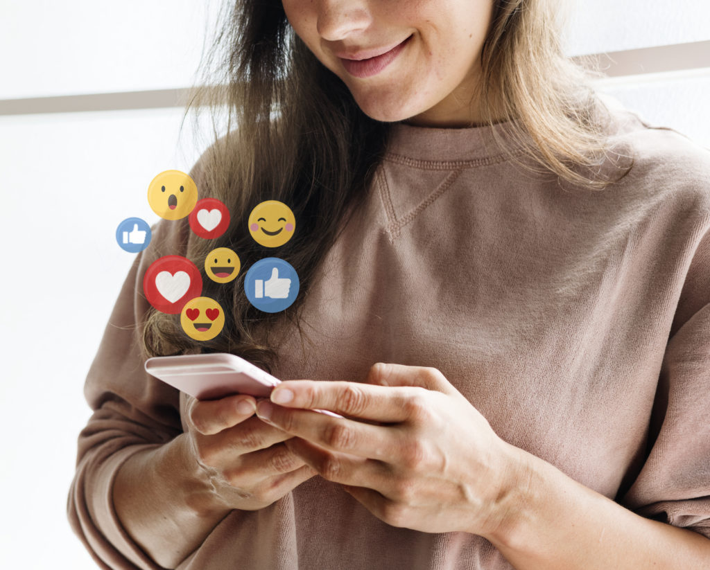 photo of a smiling young white woman looking down at her phone with overlay of animated floating Facebook likes, hearts and emoji faces emanating from the phone screen