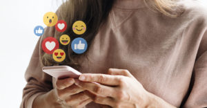 photo of a smiling young white woman looking down at her phone with overlay of animated floating Facebook likes, hearts and emoji faces emanating from the phone screen