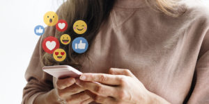 photo of a smiling young white woman looking down at her phone with overlay of animated floating Facebook likes, hearts and emoji faces emanating from the phone screen