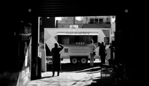 black and white photo looking down a dark alley toward a food truck bathed in light on the street with customers lined up