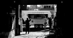 black and white photo looking down a dark alley toward a food truck bathed in light on the street with customers lined up