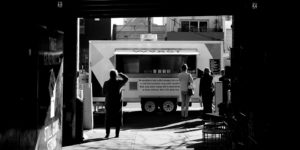 black and white photo looking down a dark alley toward a food truck bathed in light on the street with customers lined up