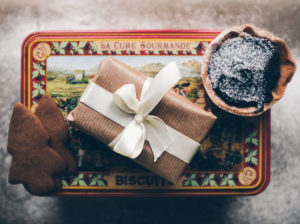 photo of a wrapped gift and cookies on top of a Christmas biscuit tin with snowy background